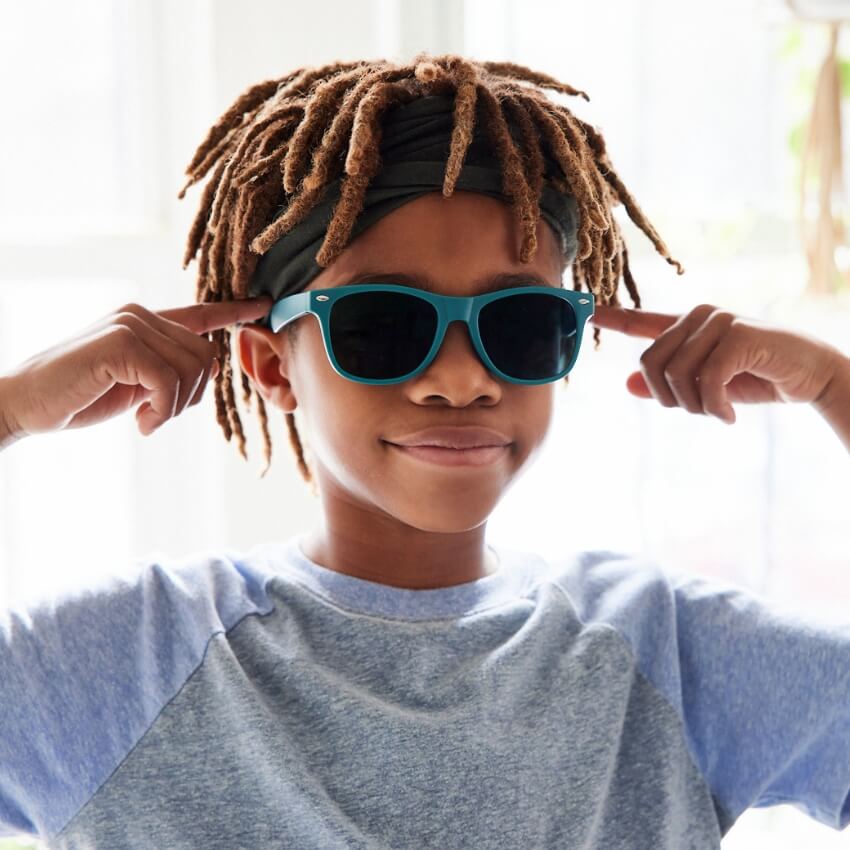 Child wearing teal sunglasses and a grey and light blue shirt, pointing towards the sunglasses' arms.