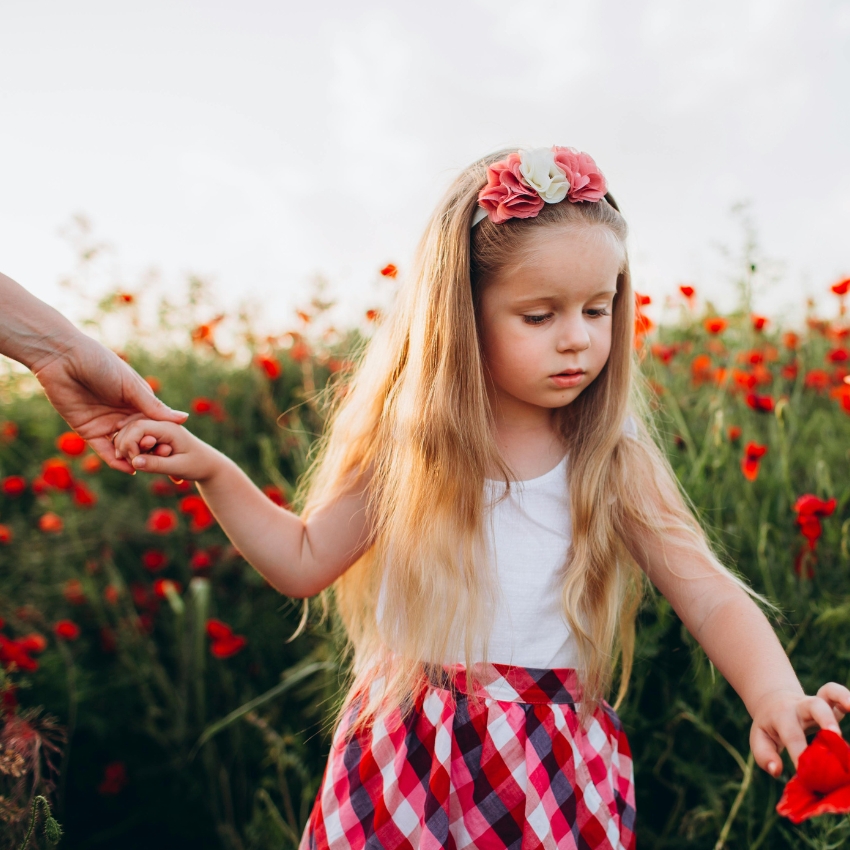 Child with long hair wearing a white top, red and white checkered skirt, and a floral headband in a flower field. Holding an adult's hand.