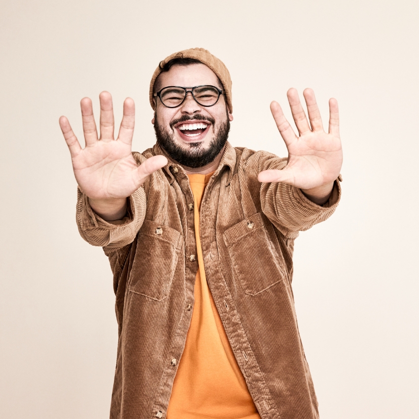 Man wearing a brown corduroy jacket, orange shirt, beanie, and glasses, smiling and raising both hands.