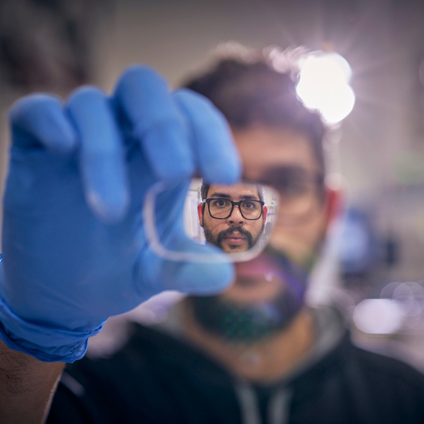 Gloved hand holding a lens with a bearded man's face in focus through the lens.