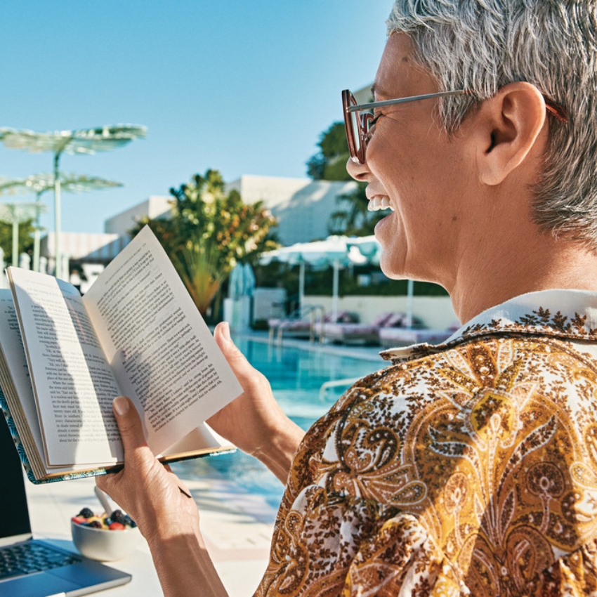 Person reading a book with both hands, paperback held open. Bowl of fruit and laptop on a table nearby.