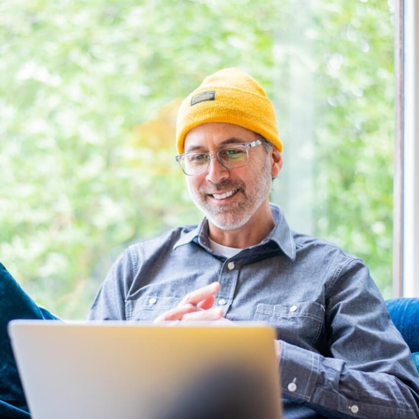 Smiling man wearing a yellow beanie hat and glasses, working on a laptop.