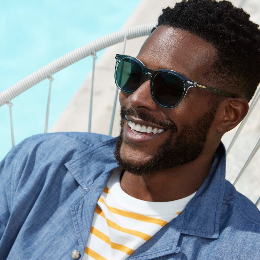 Man smiling, wearing dark green sunglasses and a blue chambray shirt over a yellow and white striped T-shirt.