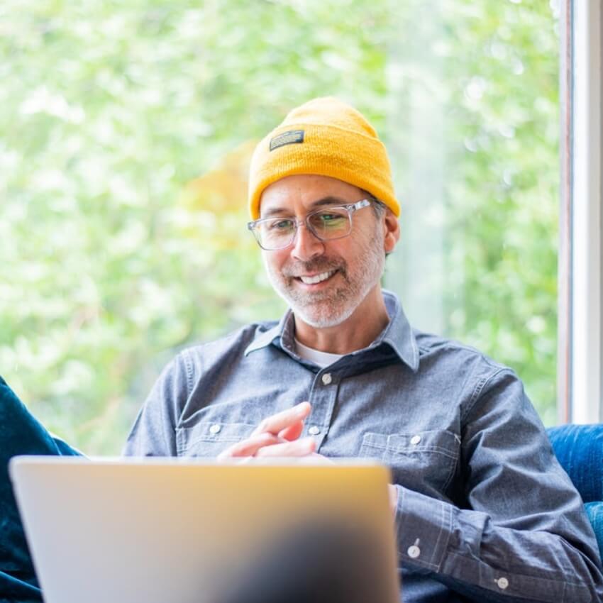 Man wearing a yellow beanie and glasses, smiling while using a laptop.