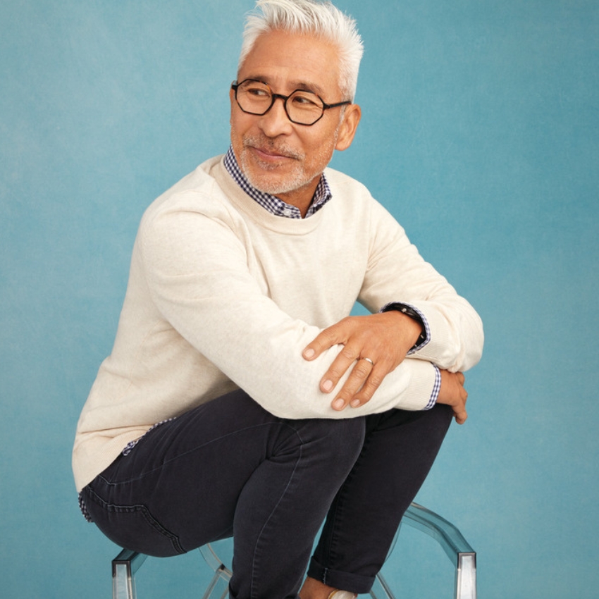 Man wearing a beige sweater, checkered shirt, and dark jeans, seated on a transparent chair.