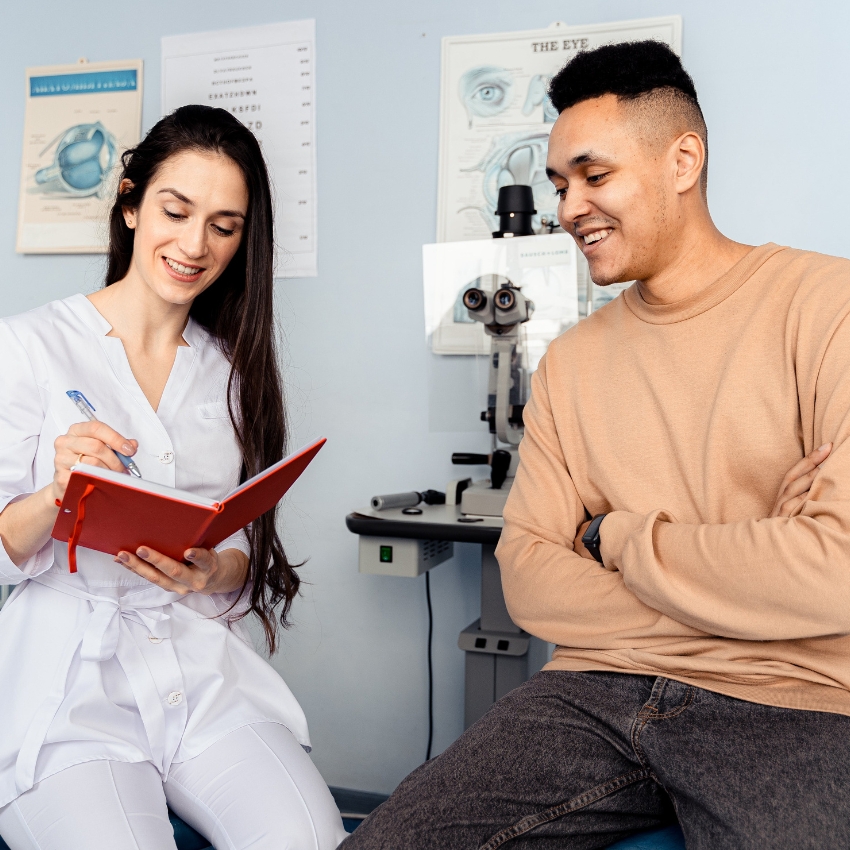 Optometrist writing notes in a red notebook while smiling at a patient. Eye examination equipment in view.