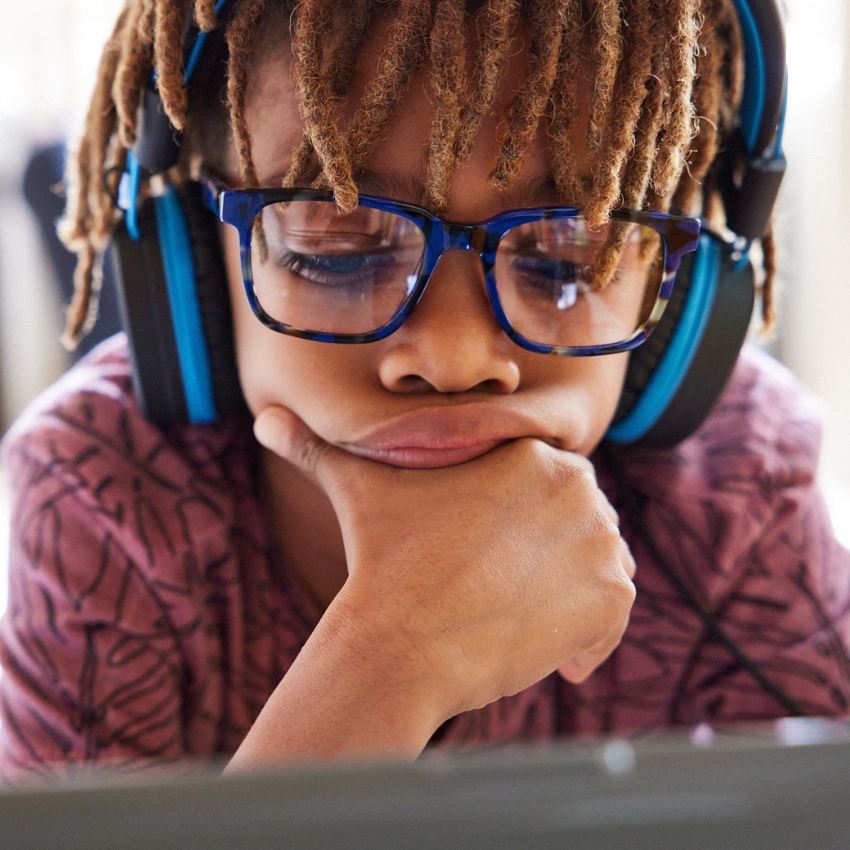 Child wearing blue-rimmed glasses and blue over-ear headphones, resting their chin on their hand.