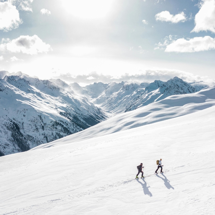 Two skiers trekking up a snowy mountain slope with snow-capped peaks in the background.