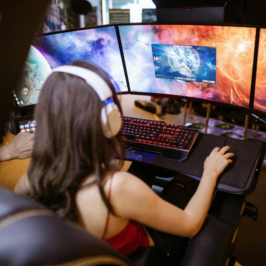 Person using a triple-monitor gaming setup with a red backlit keyboard and gaming mouse.