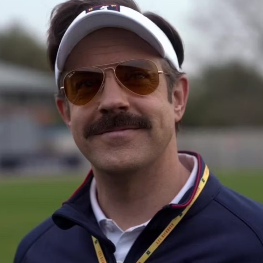 Man with a mustache wearing a white visor, aviator sunglasses, and a lanyard with a black jacket.