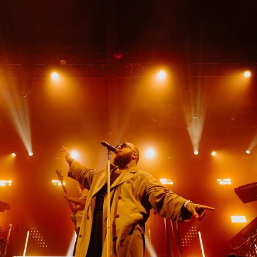 Performer in a long coat singing on stage with dramatic orange lighting.