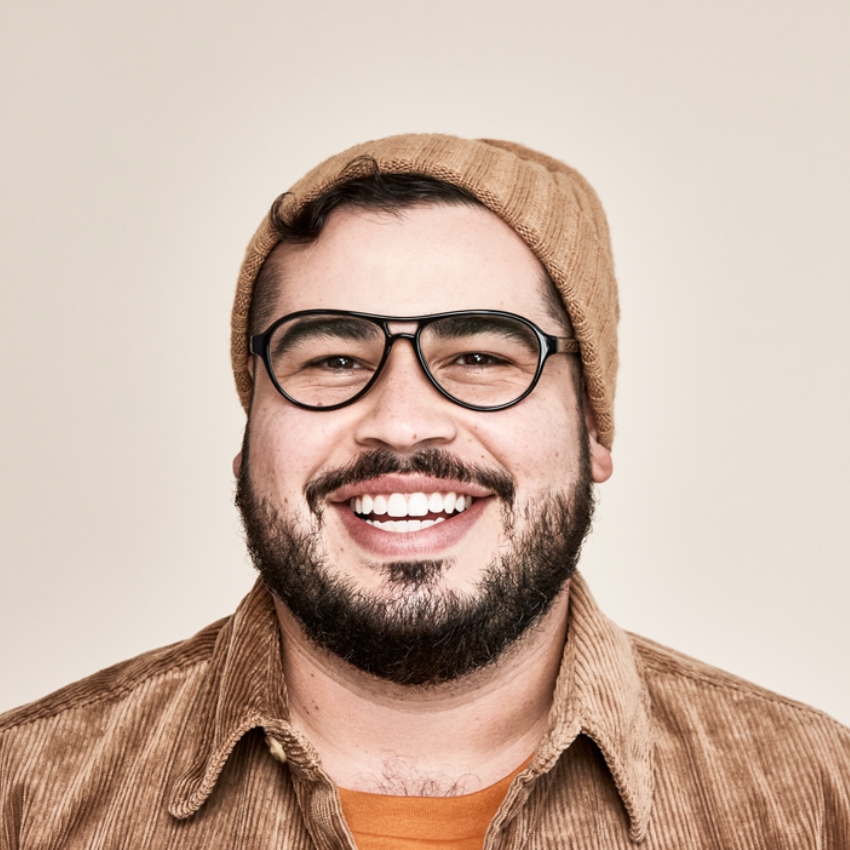 Man wearing black-rimmed eyeglasses, a brown beanie, and a light brown corduroy shirt, smiling broadly.