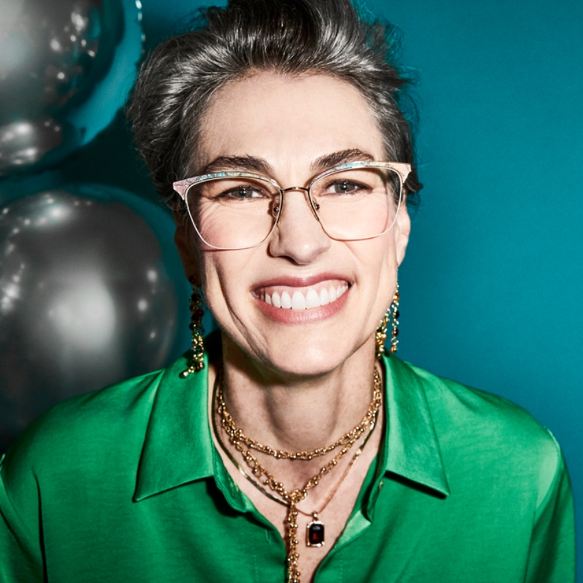 Woman wearing stylish eyeglasses, green shirt, gold necklaces, and dangling earrings, smiling broadly.