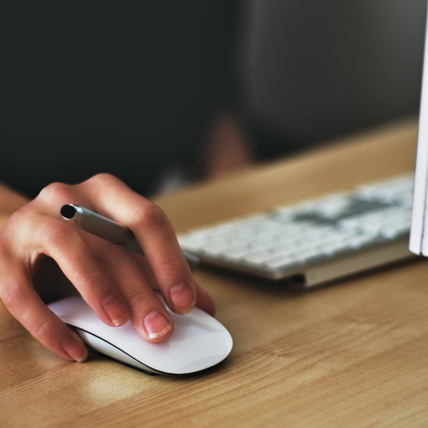 Hand holding a silver pen and a white computer mouse on a wooden desk.