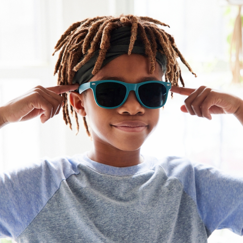 Child wearing teal sunglasses, smiling while pointing to the sides of the glasses.