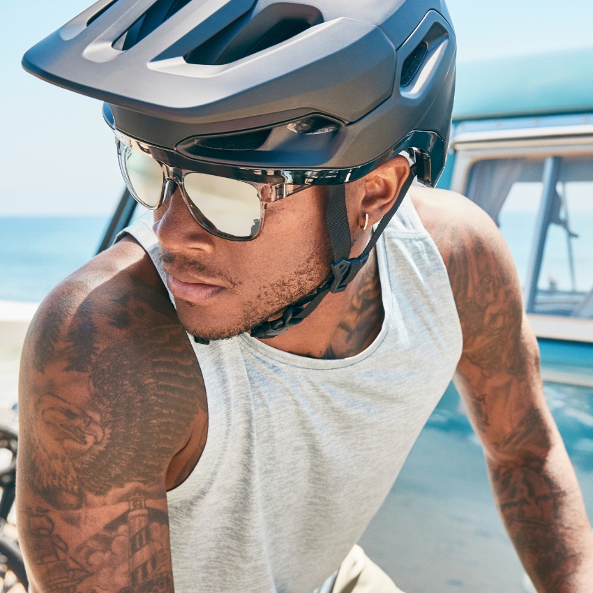 Man with sunglasses wearing a black bike helmet and gray tank top, tattoos visible on his arm.