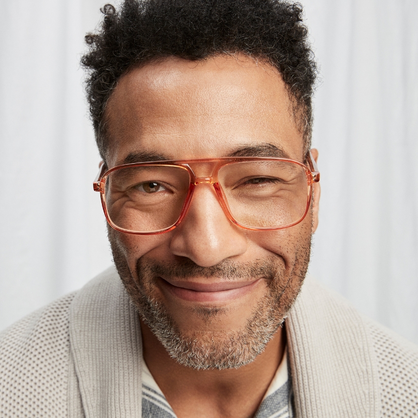 Man wearing large, translucent, orange-tinted eyeglasses and smiling with a partly closed eye.