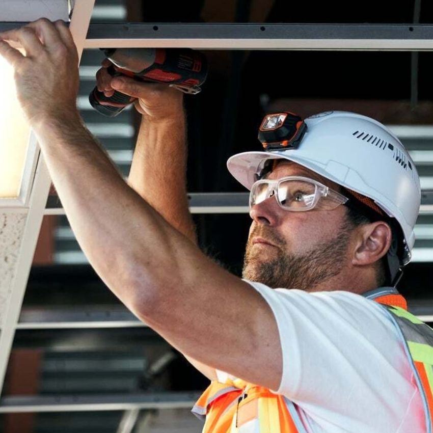 Worker in safety gear using a power drill and headlamp mount for overhead installation work.