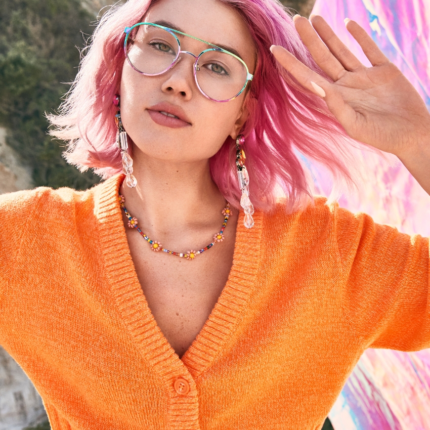 Person wearing green-rimmed glasses, an orange cardigan, beaded necklace, and crystal drop earrings.