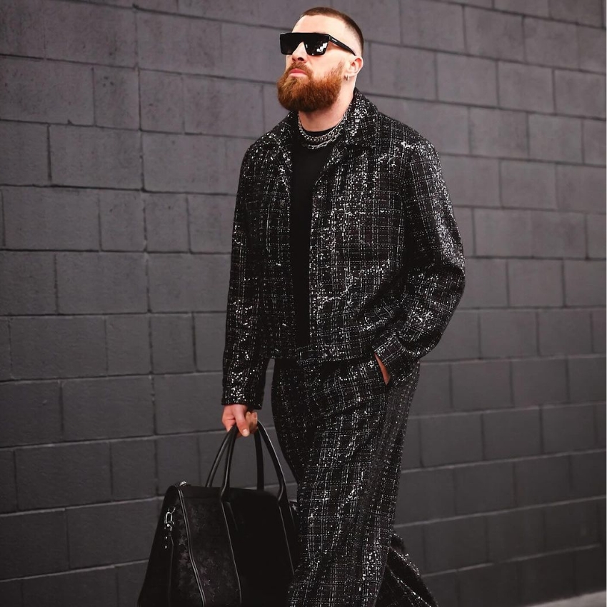 Man in a black tweed suit with sunglasses and a beard, carrying a black leather duffel bag.