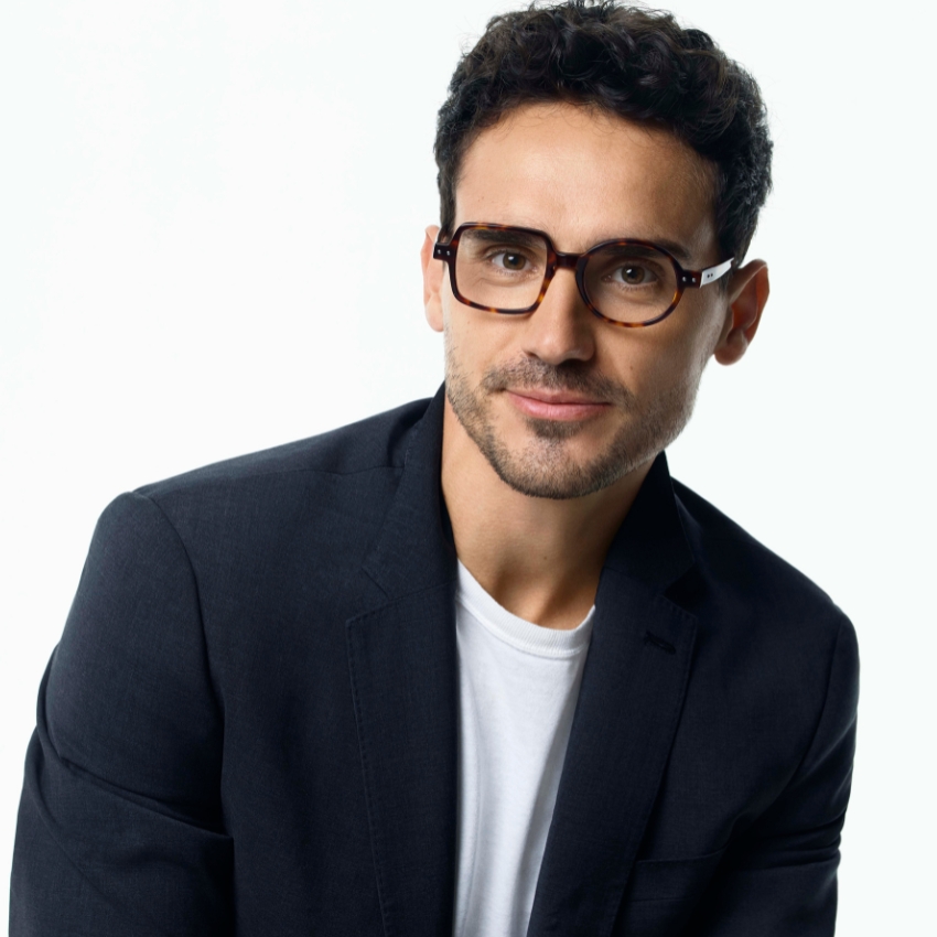 Man wearing brown round eyeglasses with a black blazer and white shirt.