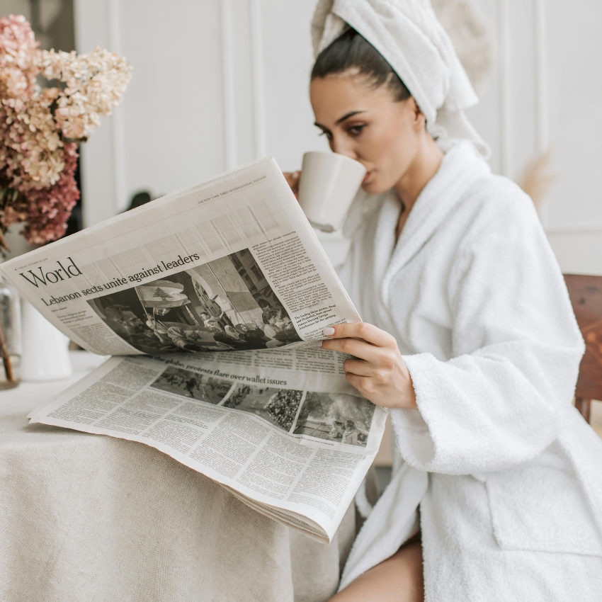 Person in white bathrobe and towel drinking from a mug while reading a newspaper. Header reads "World".