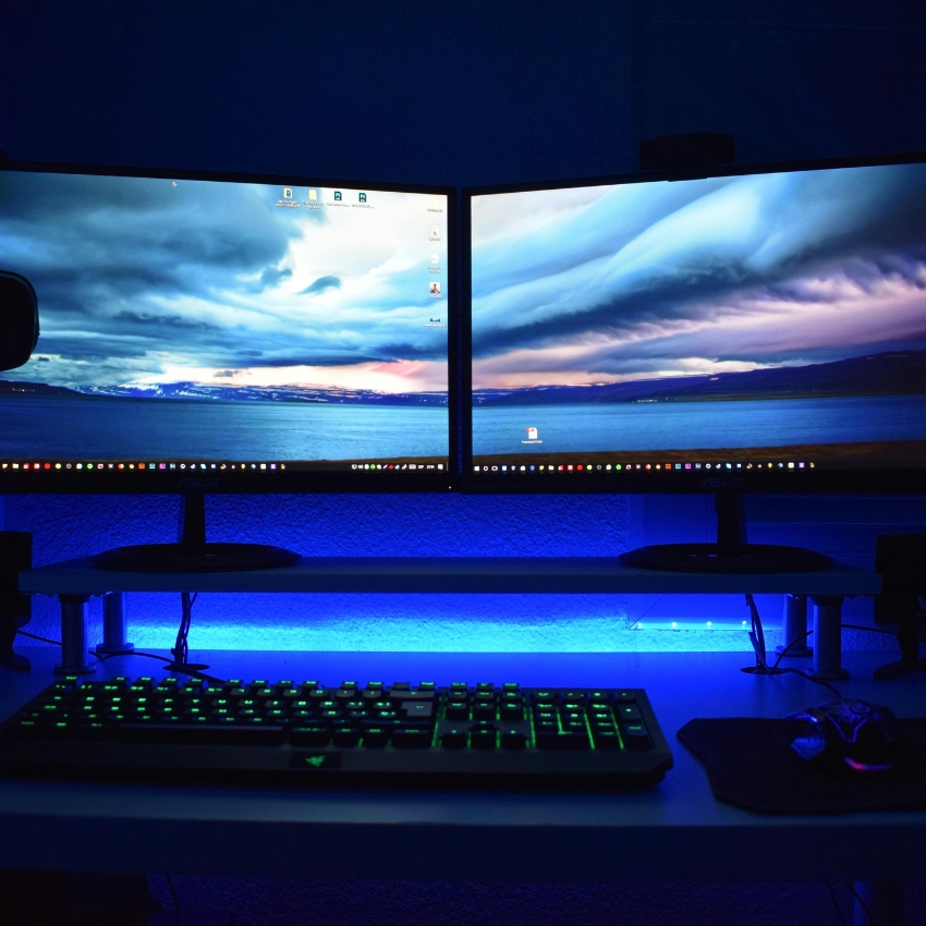 Dual monitors with a scenic desktop background, RGB backlit keyboard, and mouse on a blue-lit desk.