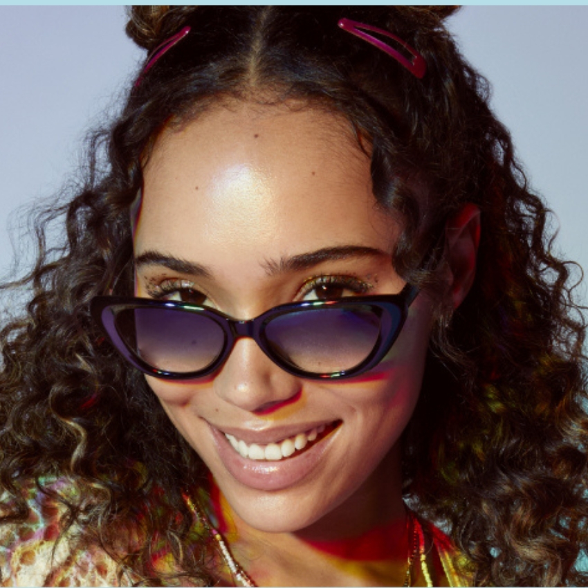 Woman wearing black cat-eye sunglasses and pink hair clips in curly hair, smiling broadly.