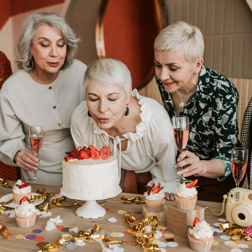 Woman blowing out candles on a cake with "60" toped with strawberries, surrounded by cupcakes.