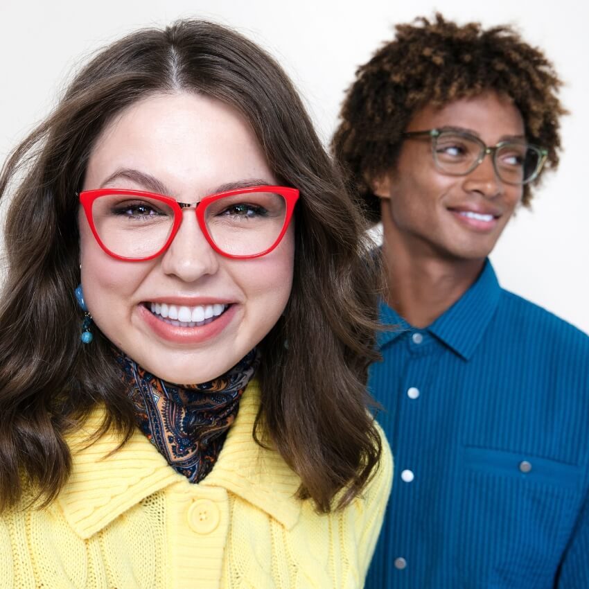 Woman wearing red cat-eye glasses and a yellow knit sweater, man in background wearing round glasses and blue shirt.