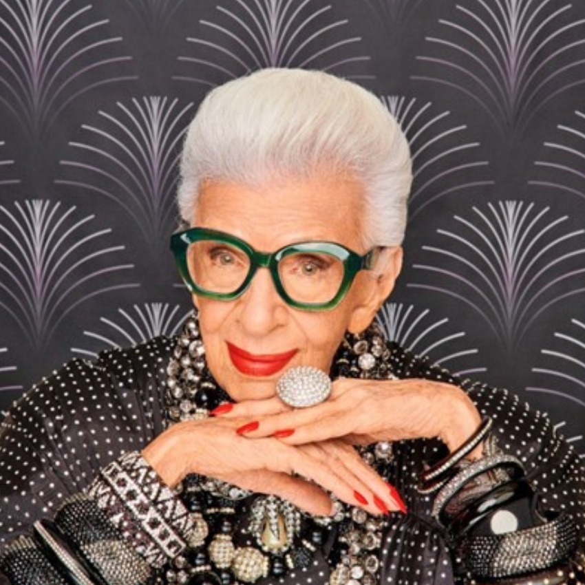 Elderly woman with short white hair, green glasses, red lipstick, and vibrant jewelry, posing confidently.