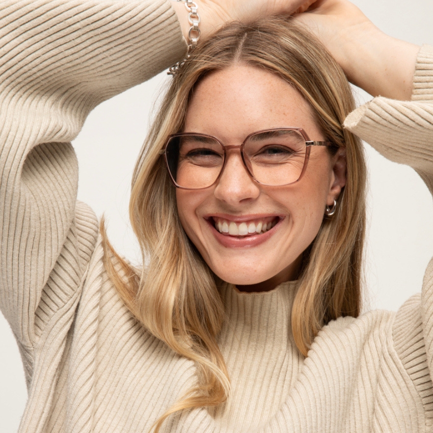 Woman wearing beige square-framed glasses and an off-white ribbed sweater, smiling with hands on her head.