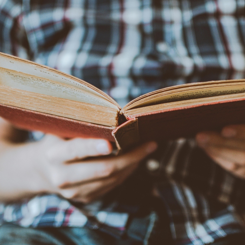 Hands holding an open vintage book with a red cover, person wearing a plaid shirt in the background.