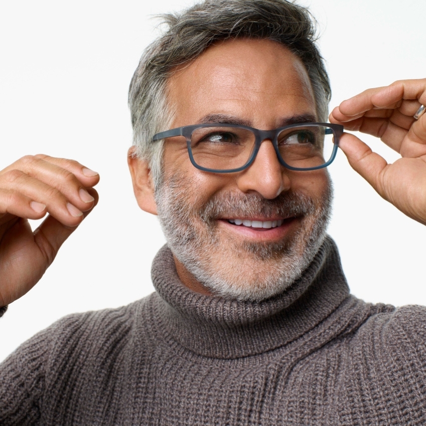 Man wearing grey-framed glasses and a grey turtleneck sweater, adjusting his glasses with both hands.