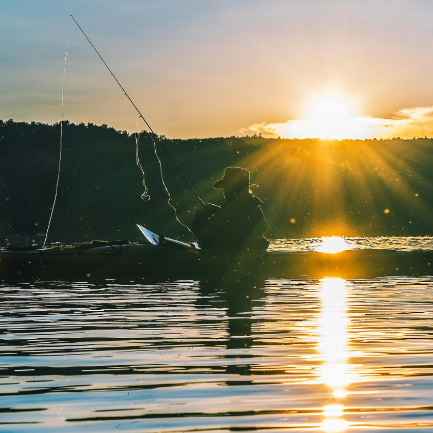 Person fishing from a kayak on a calm lake during sunset, with the sun's rays casting a golden reflection.