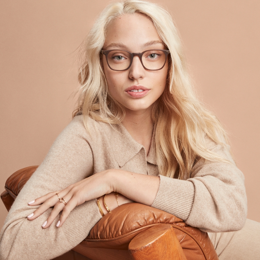 Woman wearing brown framed eyeglasses, beige sweater, leaning on a leather chair, with blonde hair.