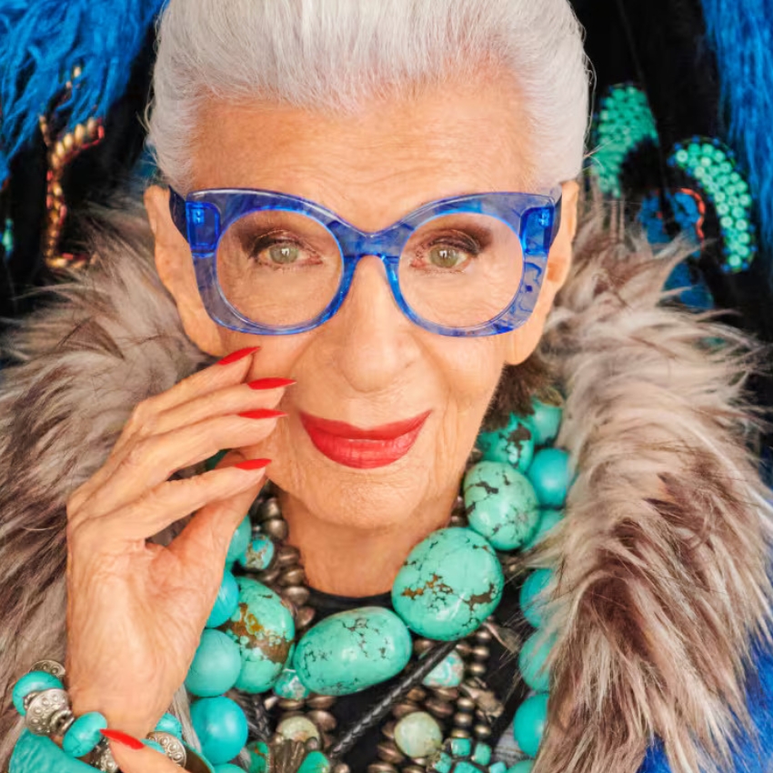 Elderly woman wearing bright blue eyeglasses and large turquoise bead necklace, with red lipstick and nails.