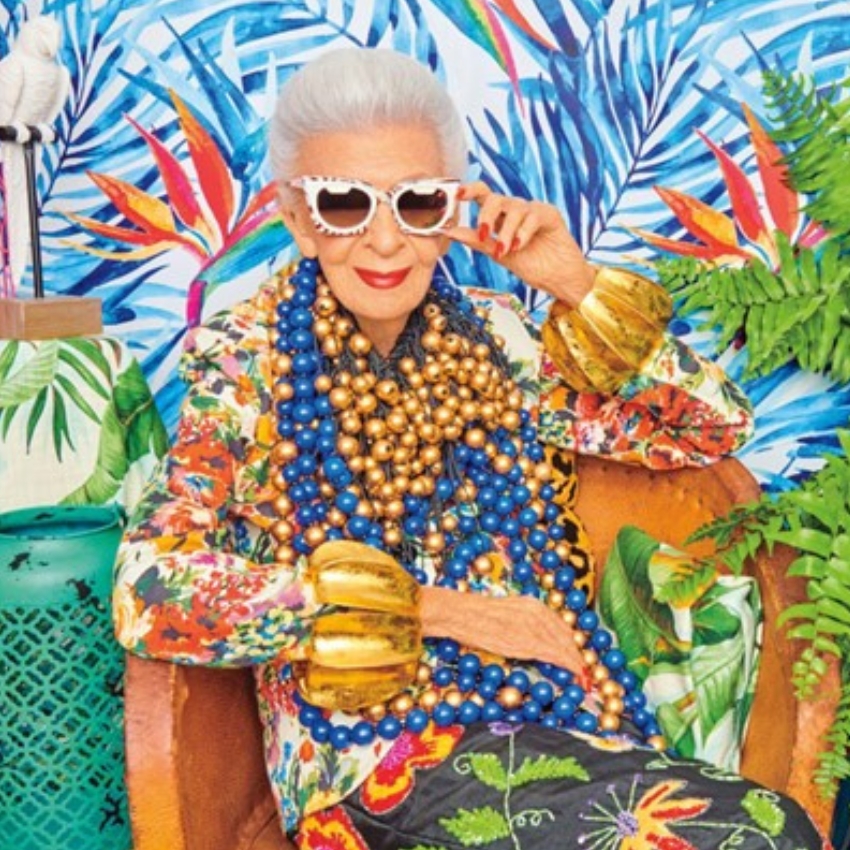 Person wearing multi-colored floral attire, gold bangles, blue and gold bead necklaces, and white sunglasses.