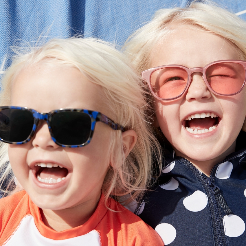 Two children smiling, one wearing blue tortoiseshell sunglasses, the other in pink transparent sunglasses.