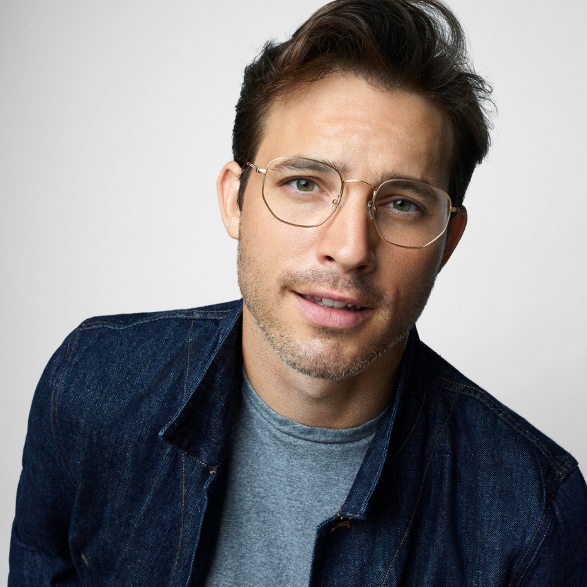 Man wearing round, gold-rimmed glasses with clear lenses. Styled in a denim jacket over a gray shirt.