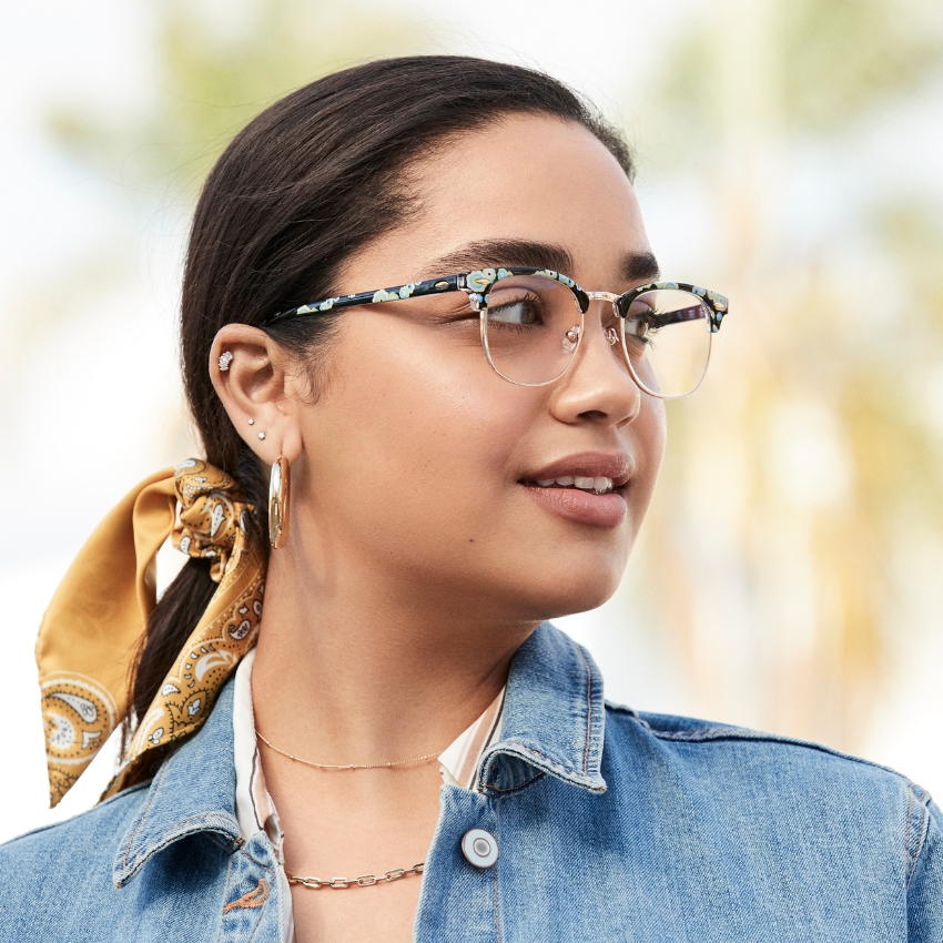 Woman wearing floral-patterned glasses with clear lenses, a denim jacket, and a yellow patterned scarf.