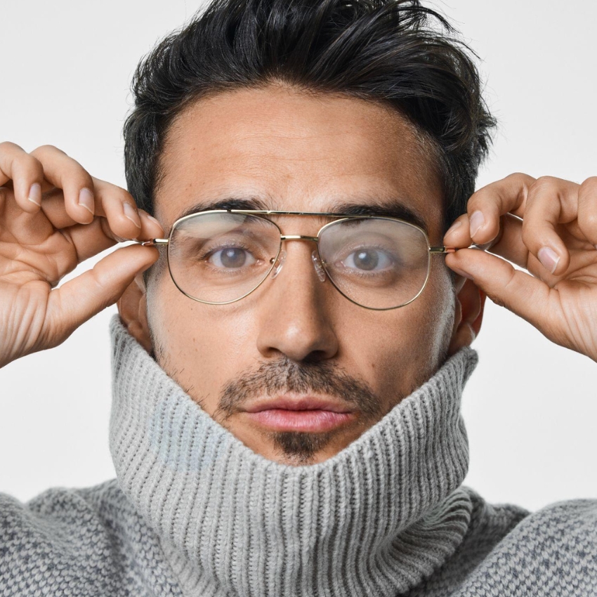 Man wearing gold-framed glasses and a gray turtleneck sweater. Hands touching the glasses frames.