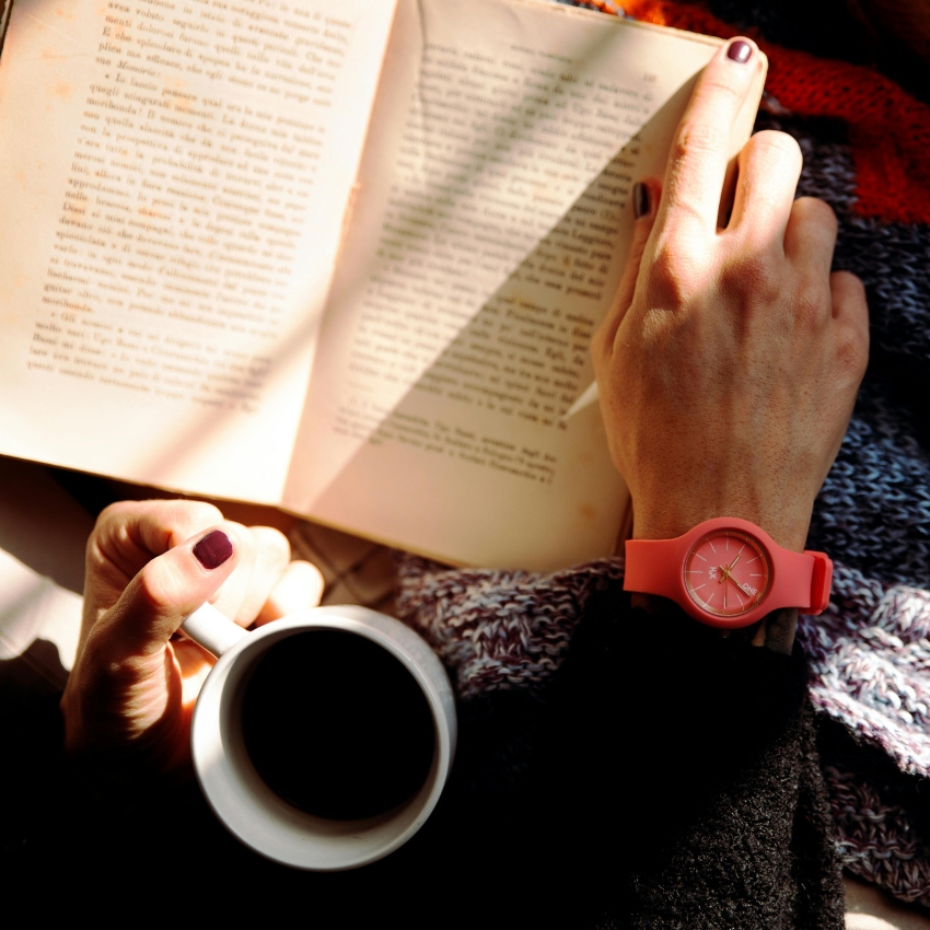 Person wearing a pink wristwatch, reading a book, and holding a cup of coffee.