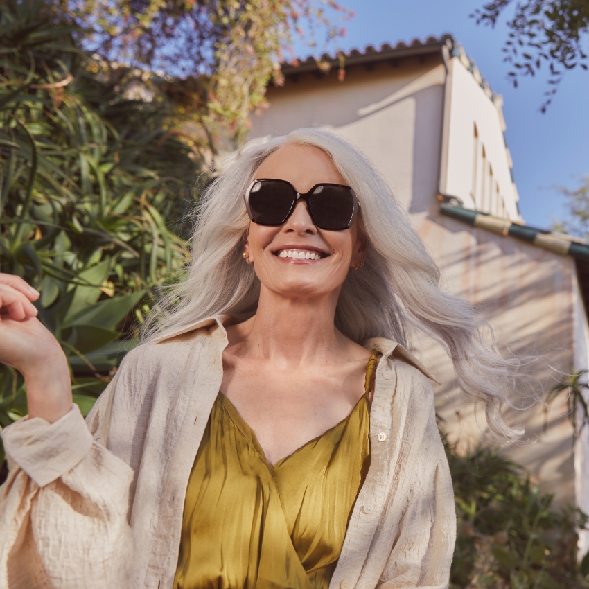 Woman wearing oversized black sunglasses and a beige shirt over a mustard dress, smiling outdoors.