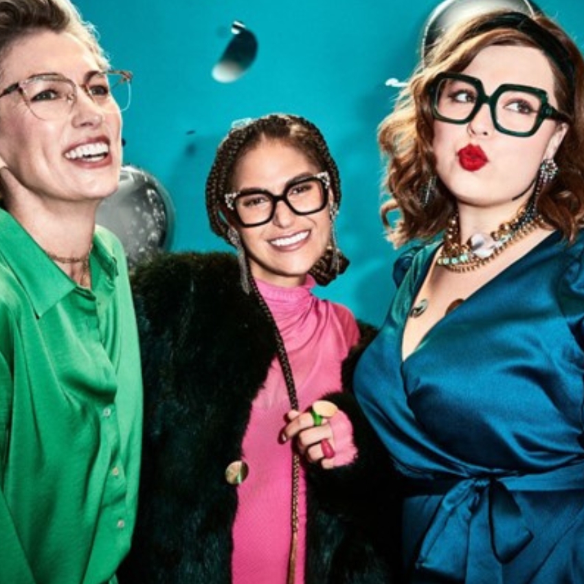 Three women wearing stylish eyeglasses, dressed in vibrant green, pink, and blue outfits, smiling and posing.