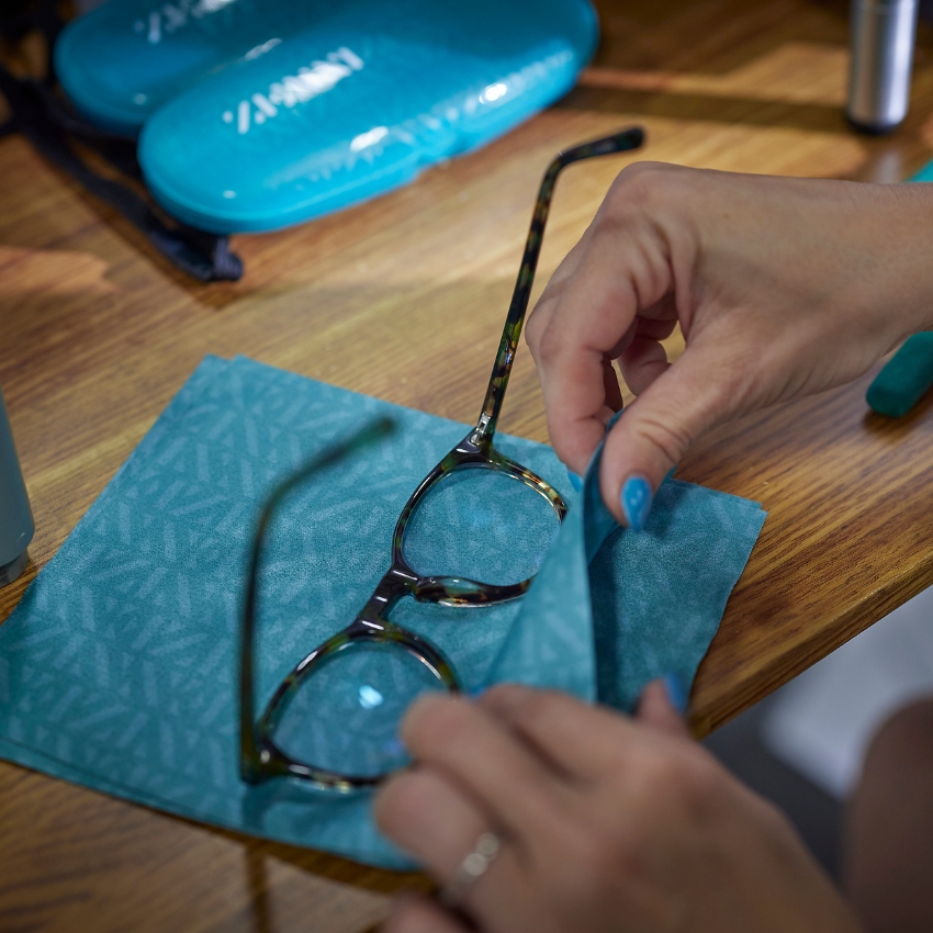 Hands cleaning eyeglasses with a teal microfiber cloth, eyeglass case in the background.