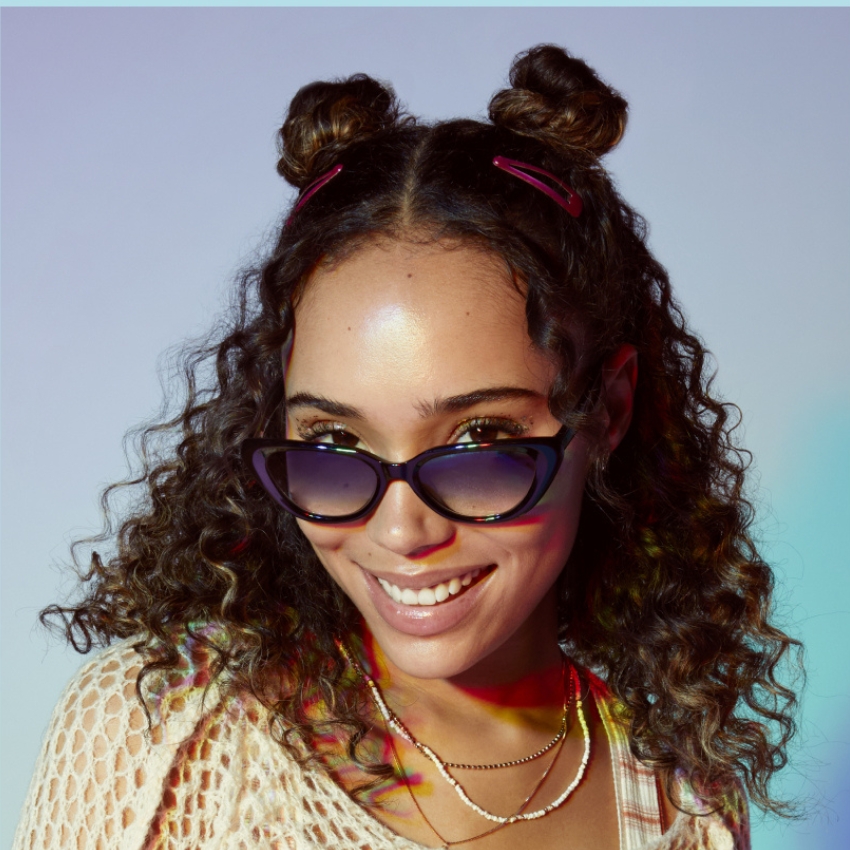 Woman wearing purple cat-eye sunglasses, two gold necklaces, and hair styled in curly buns with pink clips.