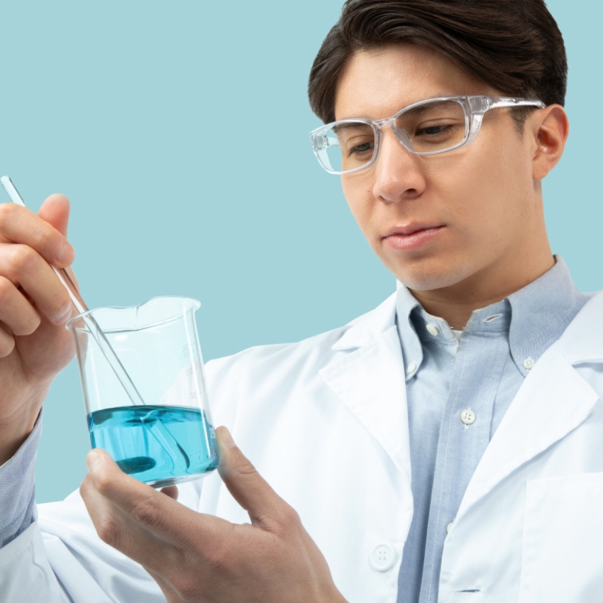 Lab technician holding a beaker with blue liquid and stirring with a glass rod, wearing safety goggles.