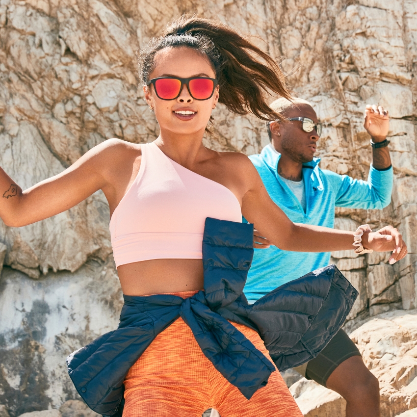 Woman in a pink one-shoulder sports bra and orange leggings with sunglasses, wearing a dark blue jacket around her waist. Man in blue athletic wear and sunglasses in the background.