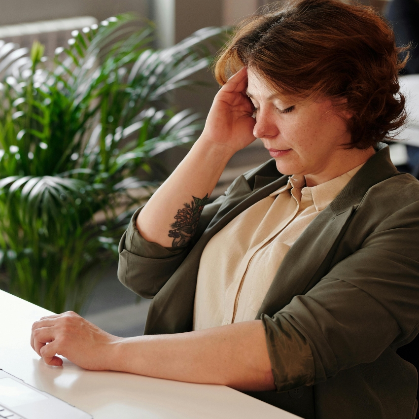 Woman in a green blazer, holding her head in one hand and looking down, sitting at a desk with a plant in the background.